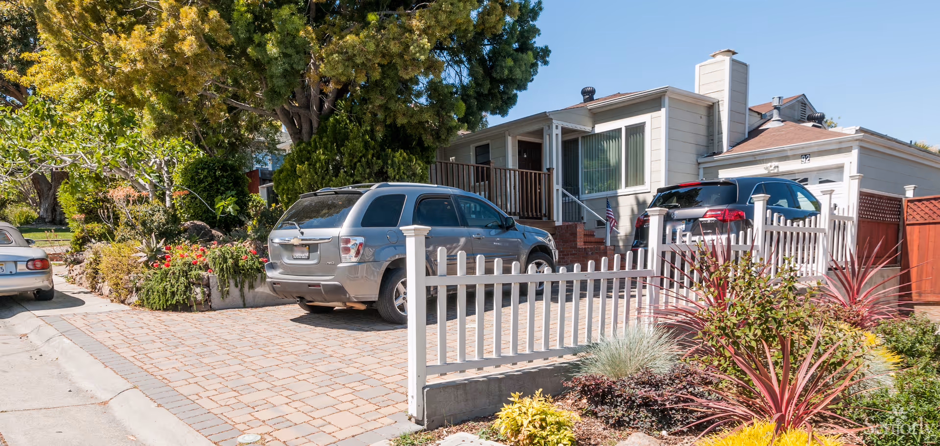 Exterior view of a single-story residential building with a driveway containing two parked cars. The house has beige siding, a small porch with stairs, and an American flag near the entrance. The front yard features a white picket fence and landscaped plants and shrubs.