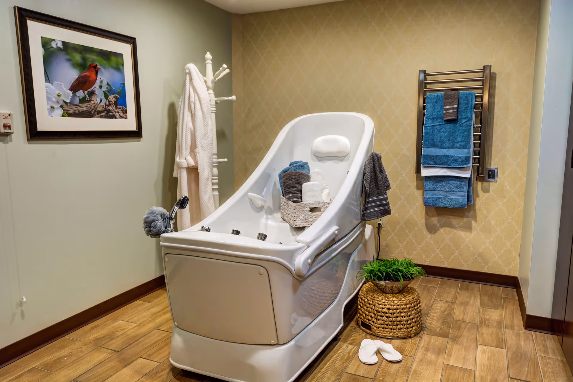 A modern white bathtub with a built-in headrest in a bathroom. The tub has a basket with rolled towels inside. There is a white robe hanging on a white coat rack, a framed picture of a red bird on the wall, a towel rack with blue and gray towels, a woven basket with a green plant, and a pair of white slippers on the wooden floor.