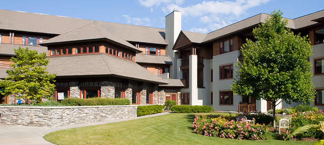 Exterior view of a multi-story senior living facility with stone and beige walls, surrounded by green grass, bushes, flowering plants, and a tree under a partly cloudy sky.