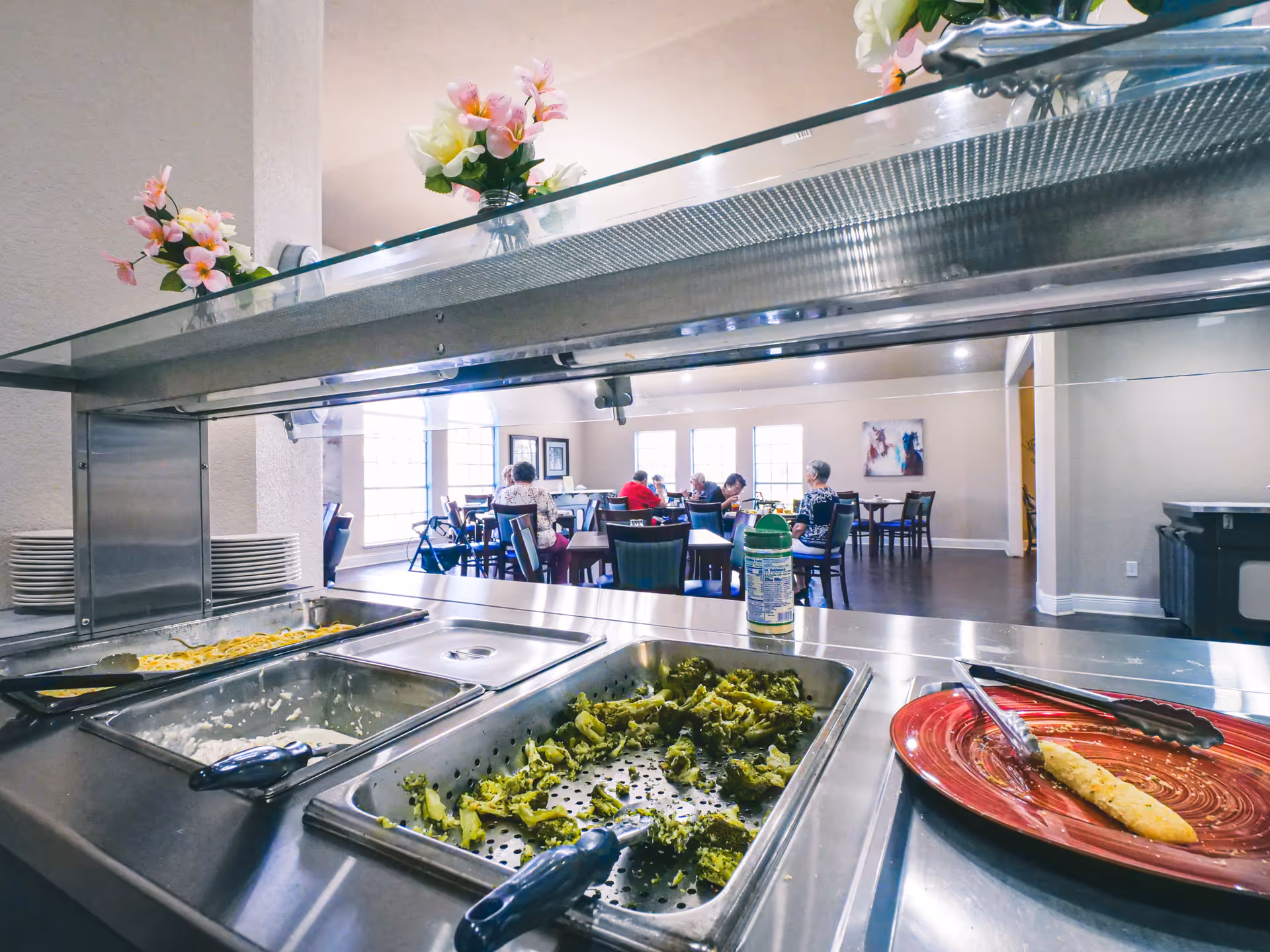 Buffet-style serving line with food trays and a dining area where residents sit at tables in the background.