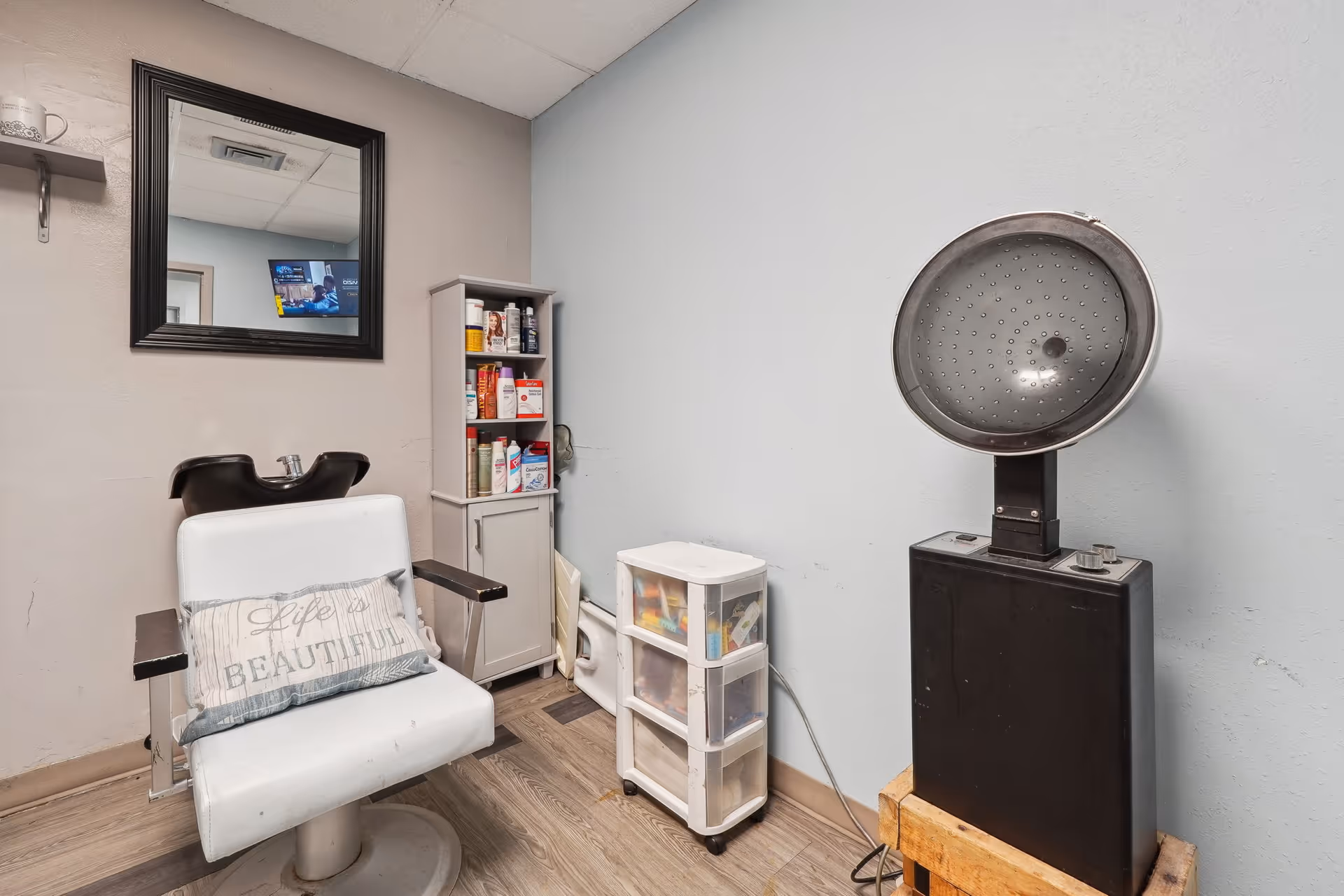 A small salon area with a white salon chair that has a pillow reading 'Life is Beautiful', a black hair washing sink behind the chair, a black-framed mirror on the wall, a small cabinet with hair care products, a plastic drawer unit, and a vintage hair dryer on a wooden stand.
