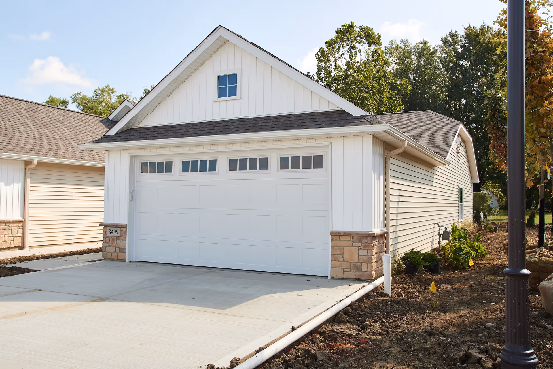 Exterior view of a white garage with a closed garage door, stone accents on the lower corners, and a small window above the door. The driveway is concrete and there are trees and plants around the building.