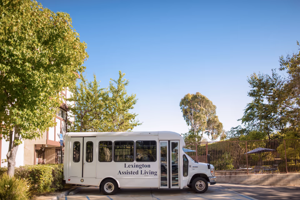 A white shuttle bus parked in a lot with 'Lexington Assisted Living' written on its side. The bus is surrounded by trees and bushes, with a clear blue sky overhead.
