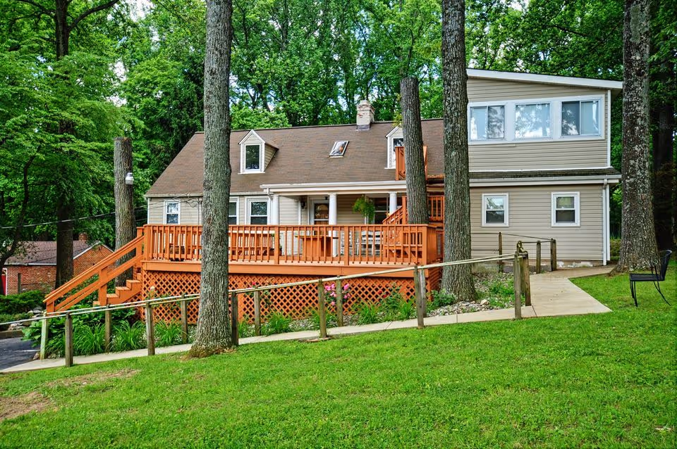 A two-story beige house with a brown roof surrounded by tall trees and green grass. The house features a wooden deck with railings and stairs leading down to a paved walkway with a handrail. There is a black chair on the right side near the walkway.