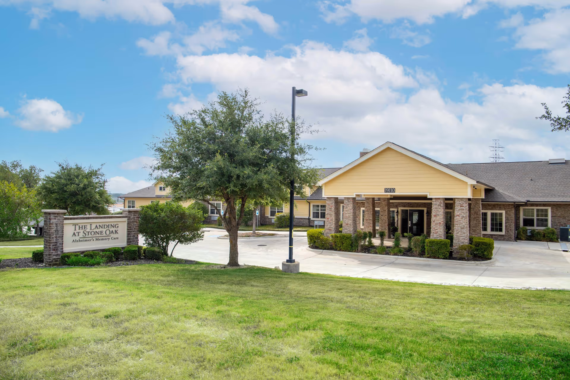Exterior view of The Landing at Stone Oak Memory Care facility showing a single-story building with a covered entrance, surrounded by green grass, trees, and a clear blue sky with some clouds.