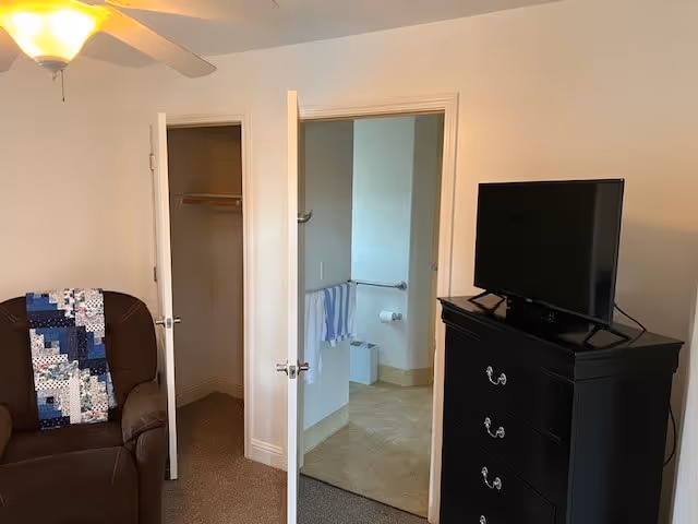 Interior view of a room in an assisted living facility showing a brown armchair with a blue and white quilt draped over it, an open closet door, an open bathroom door with a towel hanging on a rail, and a black dresser with a flat-screen TV on top.