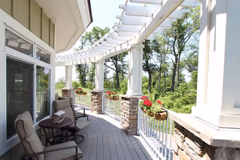A bright outdoor balcony area with cushioned chairs and a small round table. The balcony has white railings with hanging flower baskets containing red flowers, stone pillars, and a white pergola overhead. Trees and greenery are visible in the background.