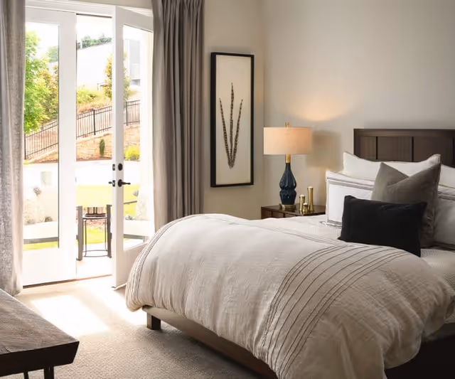 A bright bedroom with a large bed featuring white and beige bedding and multiple pillows. There is a wooden nightstand with a blue lamp and decorative items. A framed botanical artwork hangs on the wall. French doors with curtains open to an outdoor patio area with greenery visible outside.