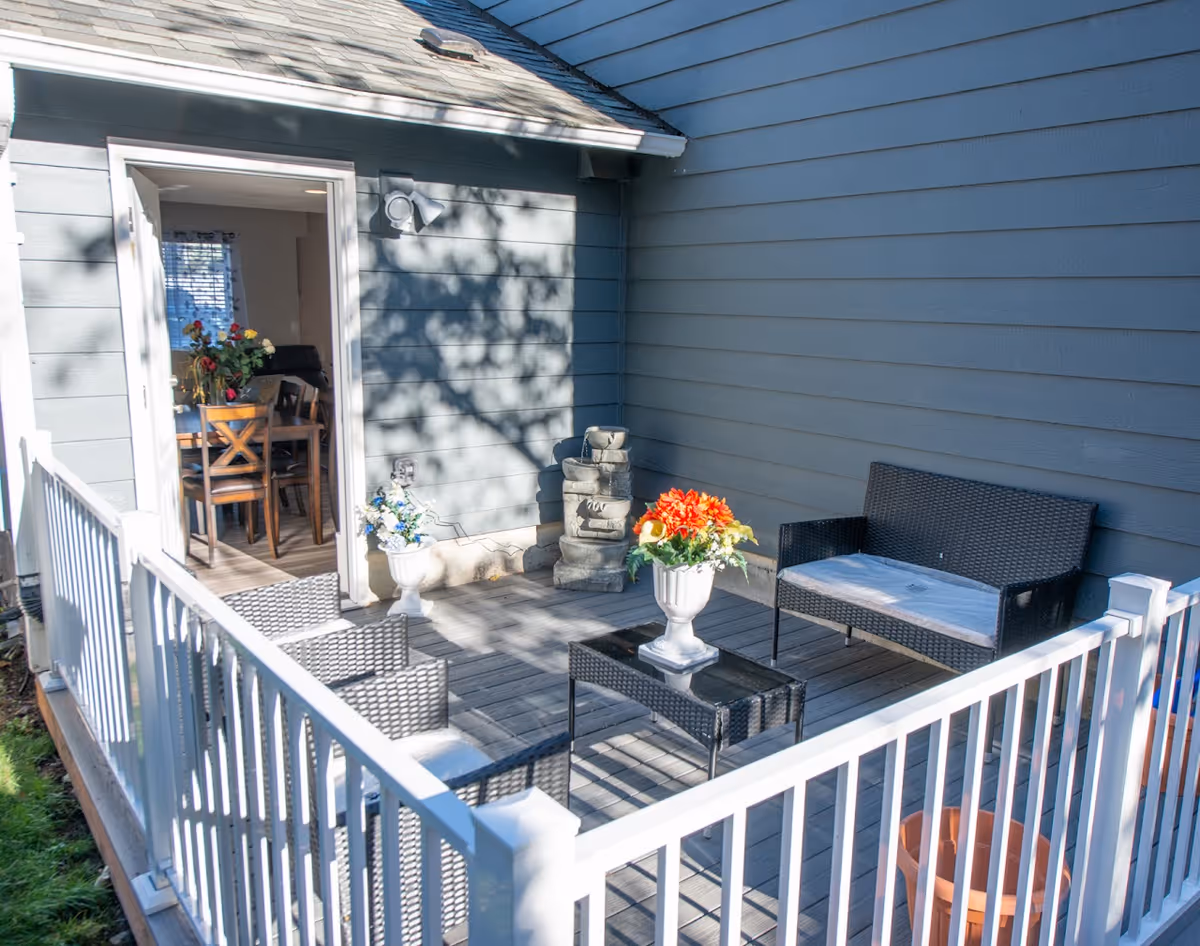 Small fenced outdoor deck with wicker seating, potted flowers, and a doorway leading into a dining area.