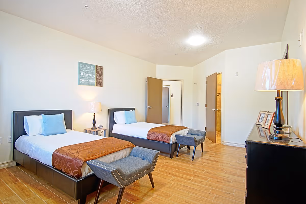 Bright shared bedroom with two twin beds, bedside lamps, footstools and a dresser on wood floors.