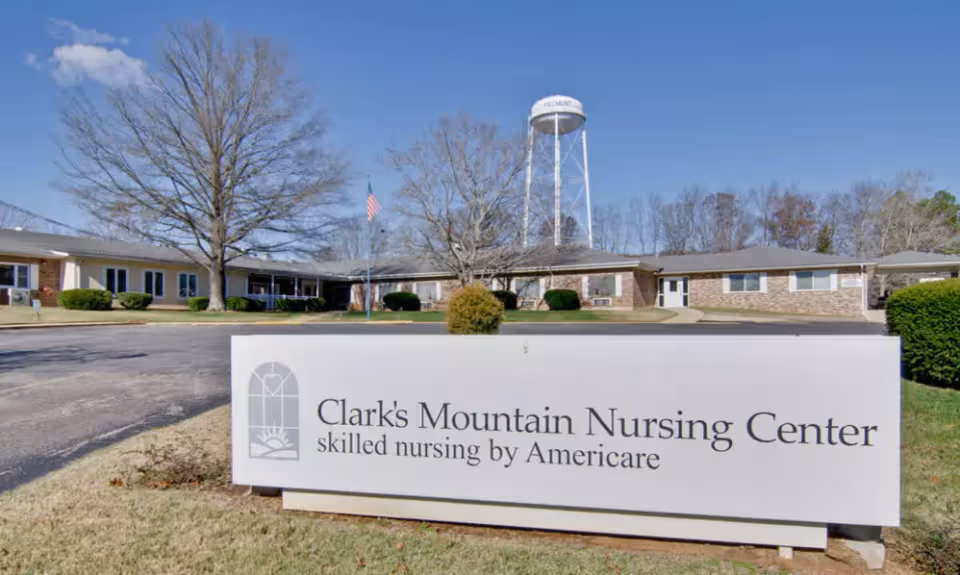 Exterior view of Clark's Mountain Nursing Center, a single-story brick building with a large sign in front that reads 'Clark's Mountain Nursing Center skilled nursing by Americare'. There are leafless trees, a flagpole with an American flag, and a water tower in the background under a clear blue sky.