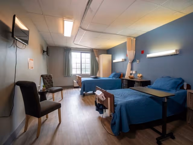 A well-lit room with two hospital-style beds covered in blue blankets, each equipped with an overbed table. There are two chairs and a small table near the wall-mounted television. The room has a large window with curtains, a cabinet, and soft lighting on the walls.