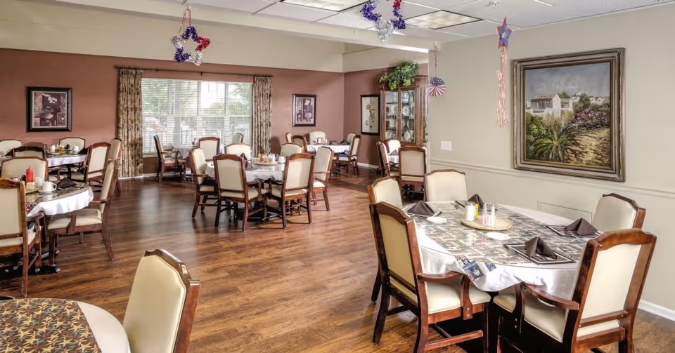 A dining room with multiple tables covered in white tablecloths and patterned runners, surrounded by wooden chairs with cream-colored cushions. The room has wood flooring, a large window with curtains, framed artwork on the walls, and patriotic star decorations hanging from the ceiling.