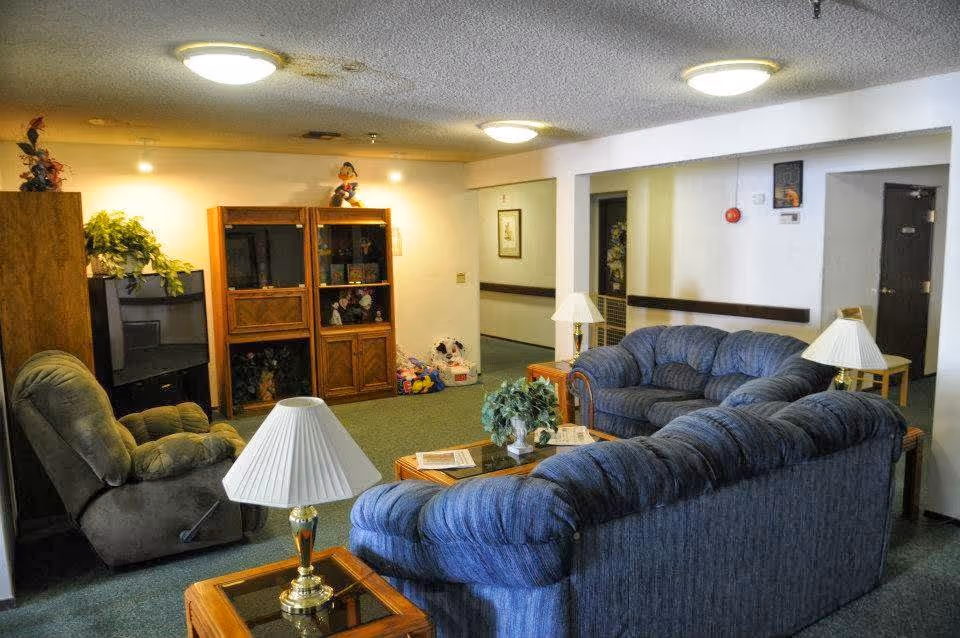 A cozy living room in a senior living facility featuring two blue upholstered sofas arranged around a glass-top coffee table with a small plant and magazines. There are two side tables with lamps, a green recliner chair, and wooden shelving units with decorative items and plants. The room has carpeted floors and white walls with ceiling lights.