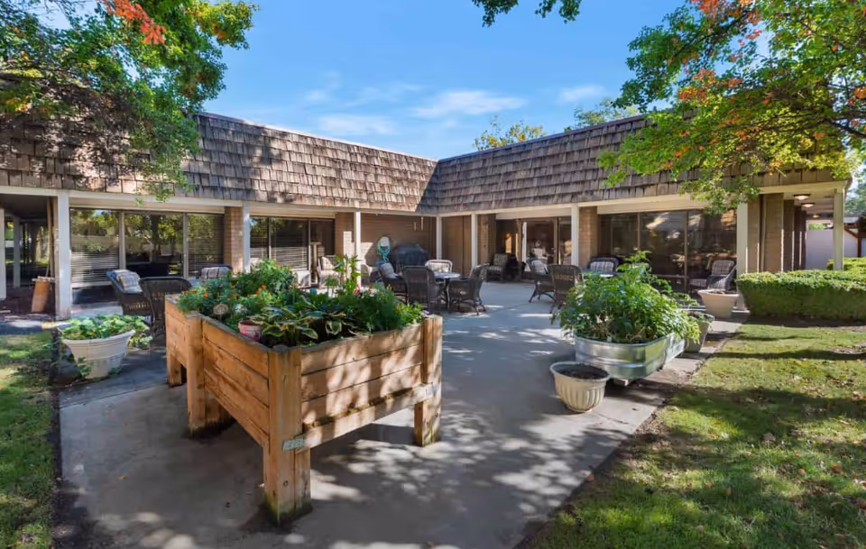 Outdoor courtyard patio at a retirement facility with raised wooden planters, potted plants and several seating areas in front of a single-story building.