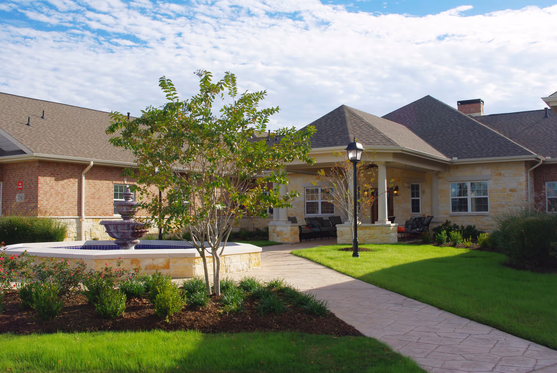 Outdoor view of Cypress Assisted Living - CyFair showing a landscaped garden with green grass, bushes, a small tree, a stone water fountain, a paved walkway, and a covered seating area attached to a building with brick and stone exterior walls under a partly cloudy sky.