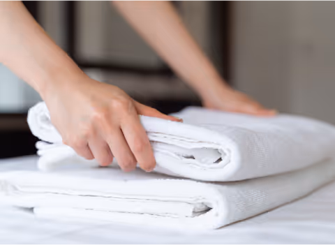 Hands fold white towels on a neatly made bed in a bedroom.