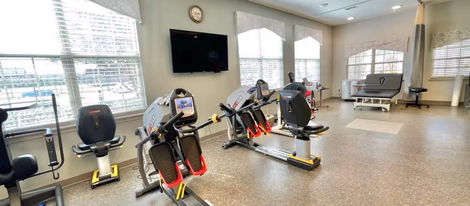 A bright exercise room with several pieces of cardio and rehabilitation equipment including recumbent bikes and leg press machines. Large windows with blinds allow natural light to fill the room. A wall-mounted TV and a clock are visible on the wall. There is also a padded examination or therapy table and a stool near the windows.