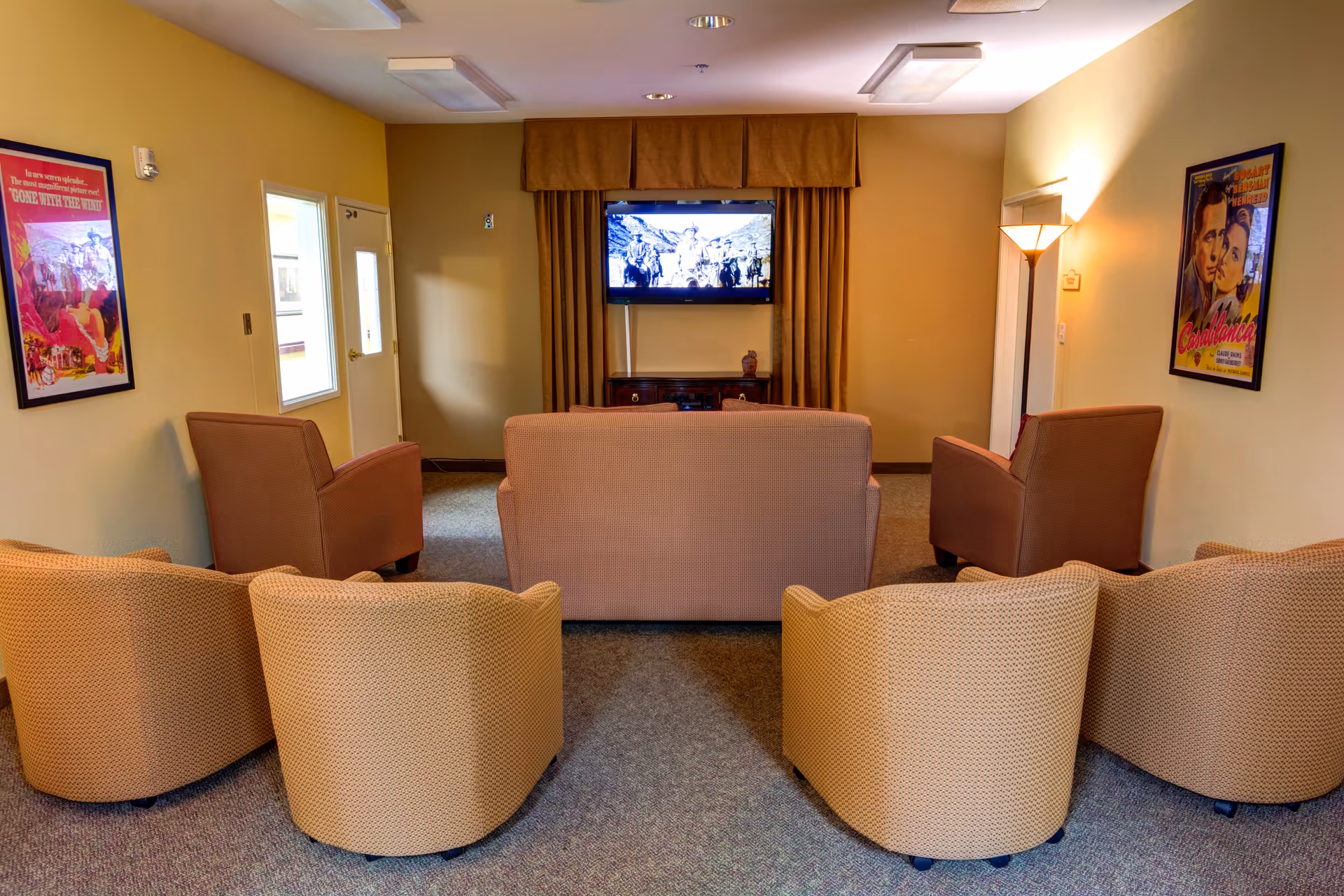 A cozy living room with a television mounted on the wall showing a black and white movie. The room has a beige couch and two brown armchairs facing the TV, with four additional beige chairs arranged in front. The walls are decorated with framed vintage movie posters, and there is a floor lamp on the right side providing warm lighting.
