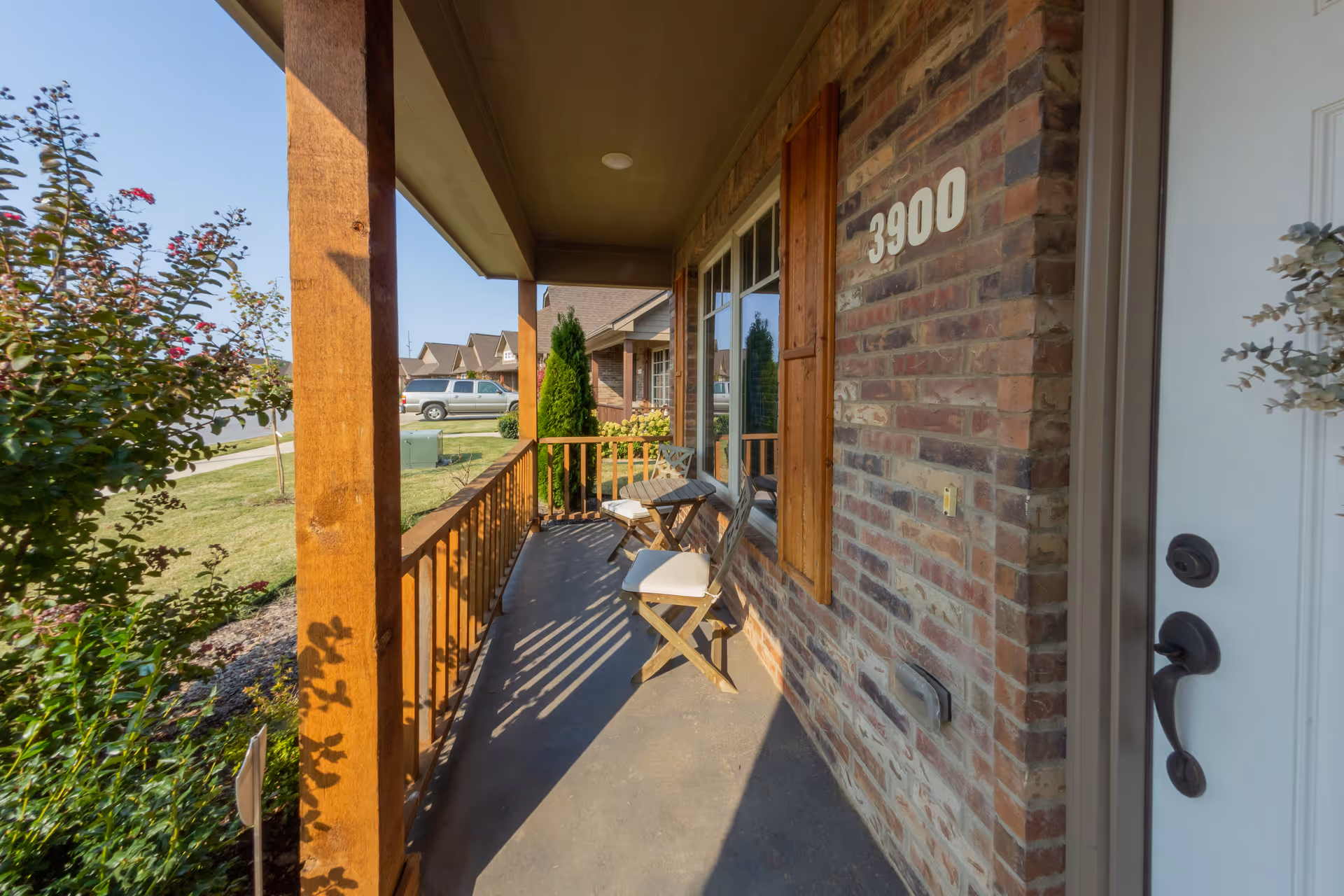 Front porch of a brick house with the number 3900 displayed on the wall. The porch has wooden railings, two wooden chairs with white cushions, and a small wooden table. There are plants and bushes near the porch, and a street with parked cars is visible in the background under a clear blue sky.