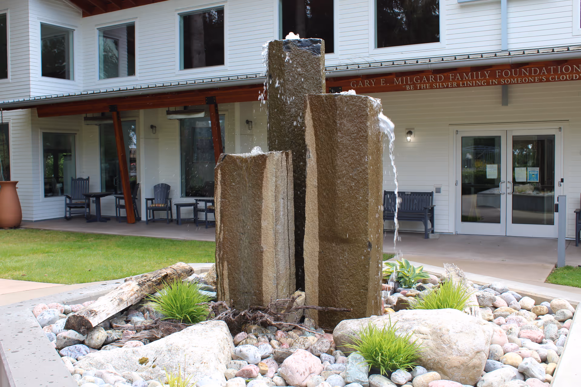 Outdoor water fountain with three vertical stone pillars surrounded by rocks and small plants in front of a white building with large windows and a covered patio area with chairs and tables.