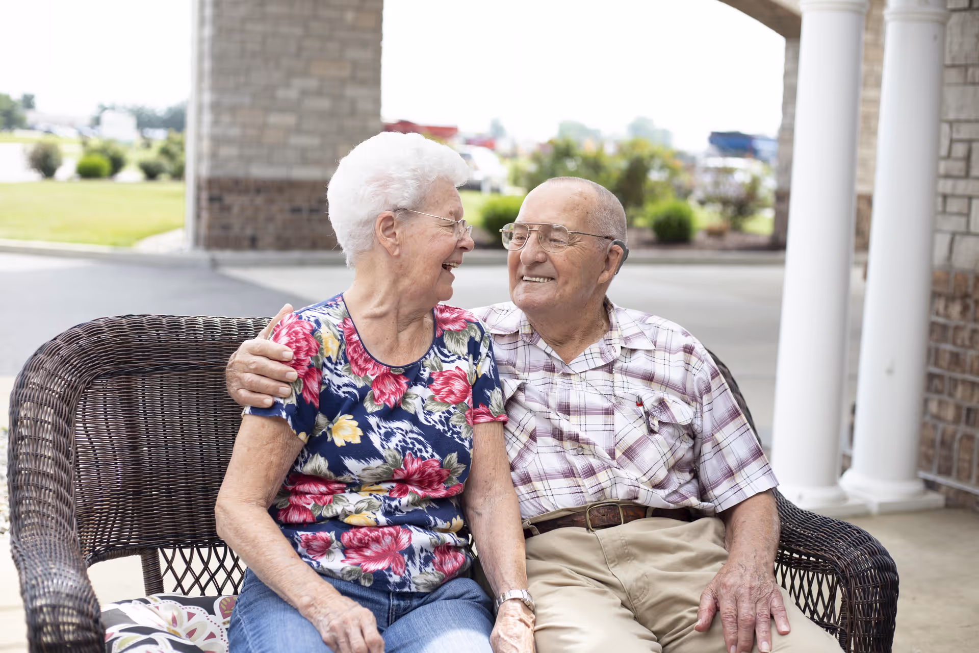An elderly couple sitting closely on a wicker bench outside a building, smiling and looking at each other affectionately. The woman is wearing a floral shirt and glasses, and the man is wearing a plaid shirt and glasses. There are green bushes and a lawn in the background.