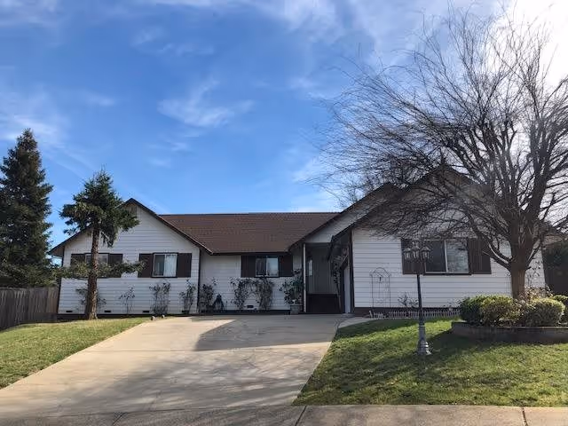 Single-story white house with brown roof and shutters, a concrete driveway, and a lawn with trees and bushes under a blue sky with some clouds.