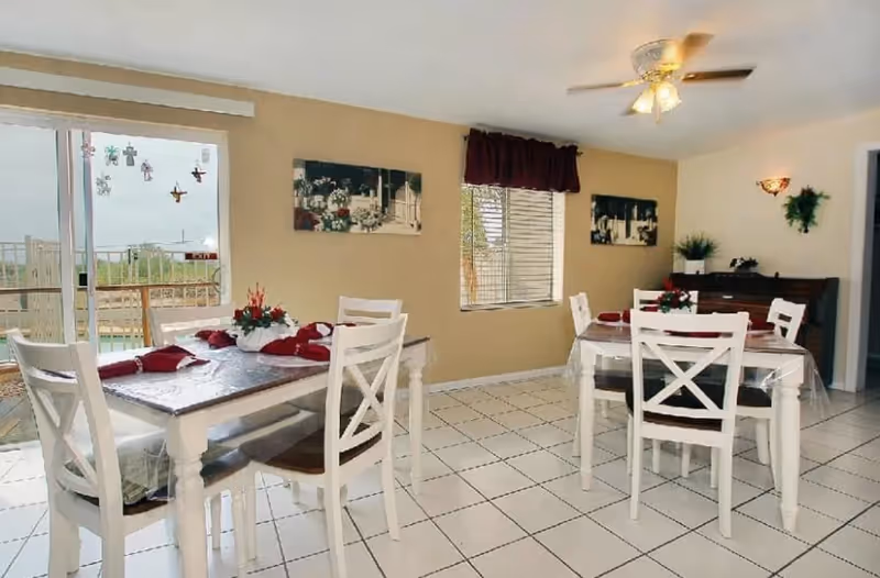 A bright dining room with two white wooden tables, each surrounded by four matching chairs. The tables are set with red napkins and floral centerpieces. The room has tiled white floors, beige walls, a ceiling fan with lights, and windows with blinds and a dark valance. There is a sliding glass door leading outside and decorative wall art and plants on the walls.