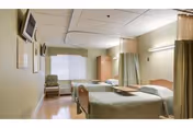 A well-lit hospital or nursing facility room with two beds separated by a curtain. Each bed has a pillow and light green bedding. There is a chair near the window at the back, a small table between the beds, and a television mounted on the wall.