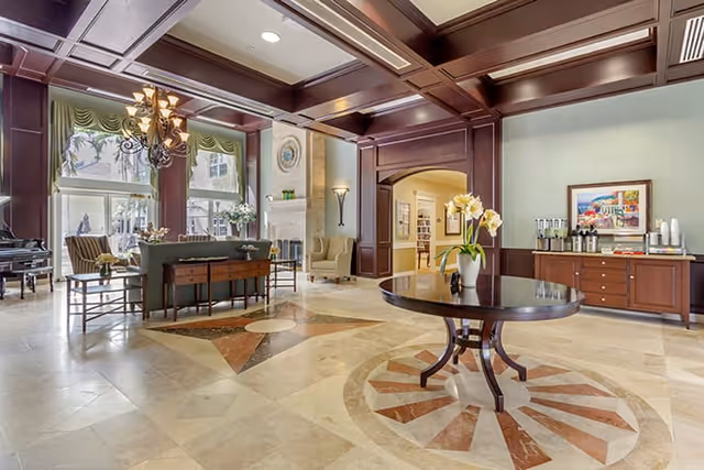 Spacious and elegant senior living facility common area with high coffered ceilings, large windows, and polished tile floors featuring decorative patterns. The room includes a round wooden table with a flower vase in the center, a seating area with chairs and a sofa, a fireplace, and a sideboard with coffee and beverage supplies. The decor is warm with wood paneling and soft lighting.