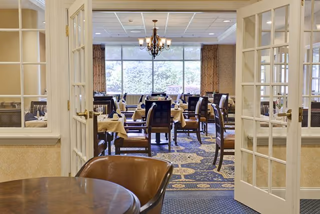 View through open French doors into a formal dining room with set tables and chairs, a chandelier, and large windows.