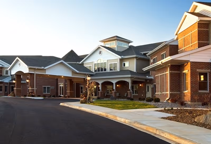 Exterior view of a senior living facility building with brick and siding, featuring a covered entrance, multiple windows, and a paved driveway with a sidewalk and landscaped areas.