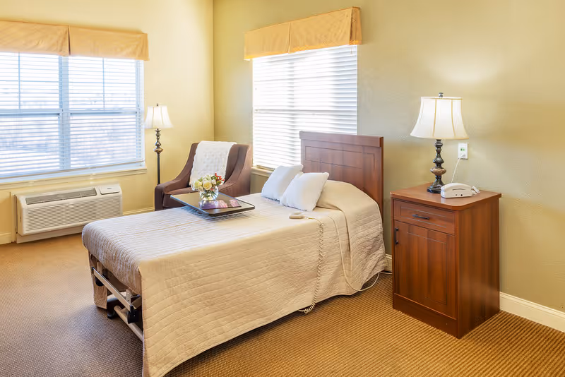 A well-lit bedroom with a neatly made bed covered in a beige quilt, two white pillows, and a tray with flowers and a book on top. Next to the bed is a wooden nightstand with a lamp and a telephone. There is a comfortable armchair with a white throw blanket near two large windows with blinds and beige valances. The room has beige walls and carpeted floor.
