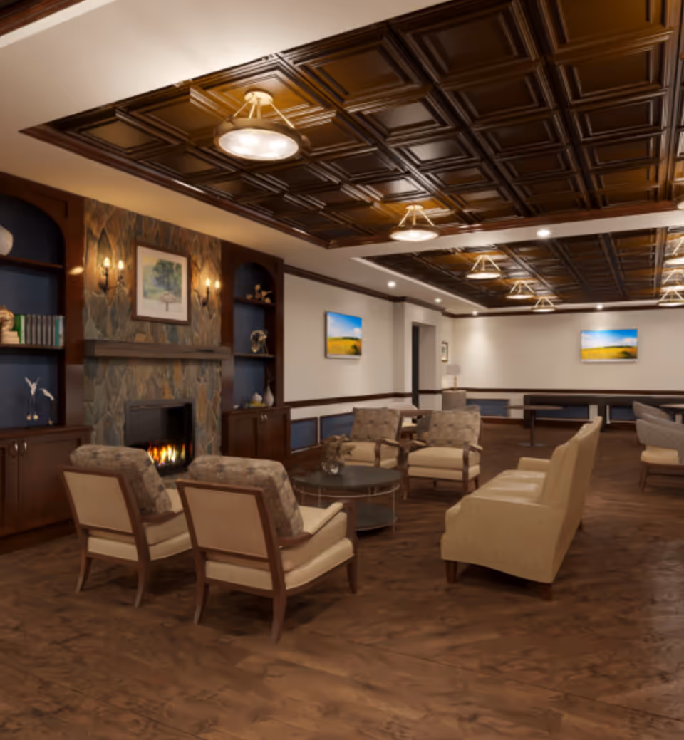 A spacious and elegant living room area with a dark wooden coffered ceiling, multiple ceiling lights, and a stone fireplace with a fire burning. The room features several upholstered armchairs and sofas arranged around a round coffee table. Built-in wooden shelves flank the fireplace, decorated with books and ornaments. The walls are adorned with framed landscape paintings, and the floor has a dark wood finish.