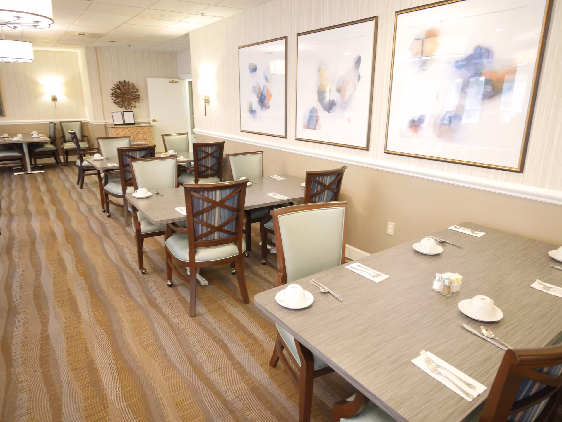 A dining room with several tables and chairs arranged neatly. Each table is set with white cups, saucers, and silverware. The room has a patterned carpet, light-colored walls with three abstract paintings, and wall-mounted lights providing illumination.