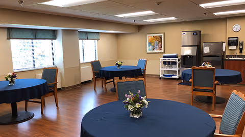 A dining room with round tables covered in dark blue tablecloths, each table decorated with a small floral centerpiece. The room has wooden flooring, several windows with green valances, and chairs with blue upholstery and wooden frames. In the background, there is a refrigerator, a water dispenser, a small cart, and a framed picture on the wall.
