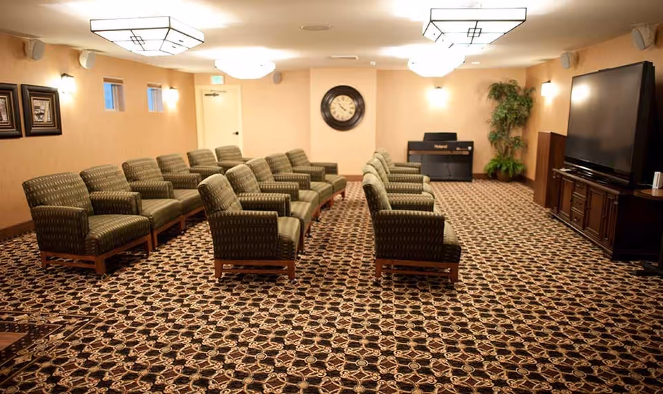 A large room with patterned carpet and two rows of green upholstered chairs facing a large flat-screen TV on a wooden cabinet. The room has beige walls, a clock on the far wall, a piano in the corner, framed pictures on one wall, and ceiling lights.