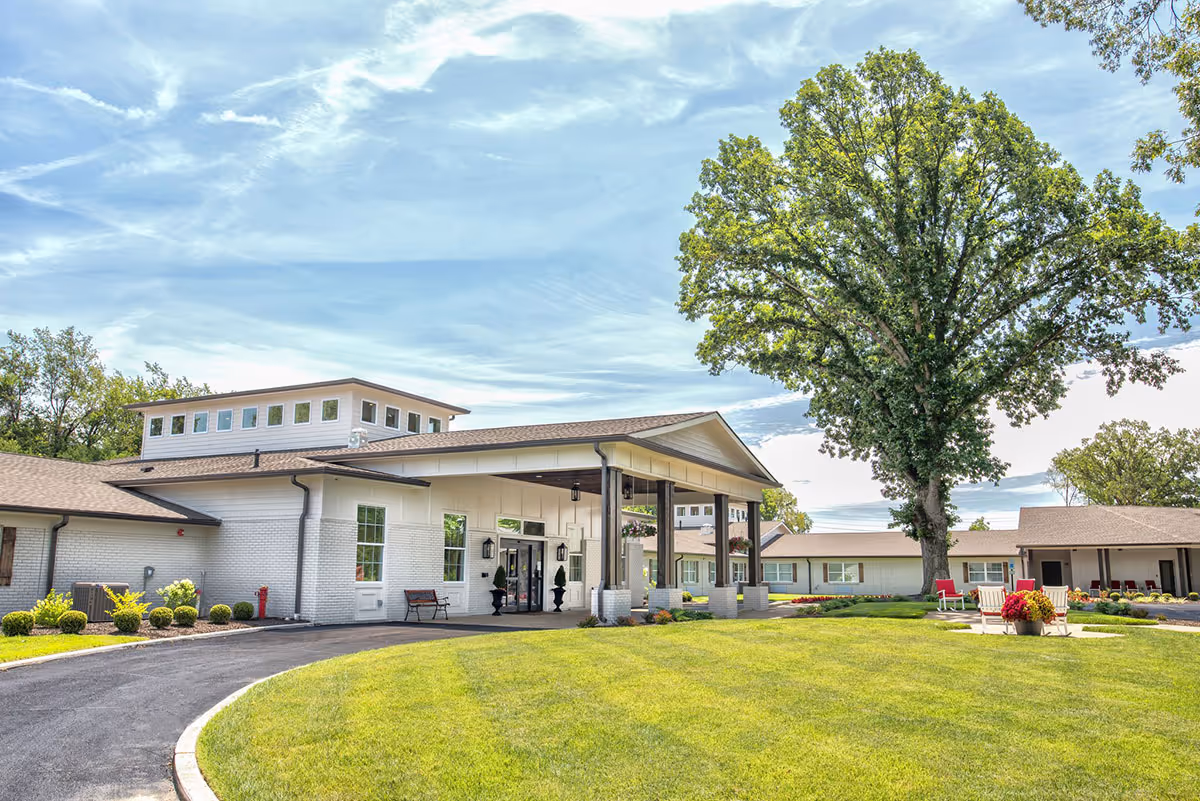 Exterior view of Dominion Senior Living At Northfield showing a single-story building with white brick walls and a covered entrance supported by wooden pillars. There is a large green lawn in front with a paved driveway curving towards the entrance. A large tree and some outdoor seating with red chairs and a flower pot are visible on the lawn under a partly cloudy sky.