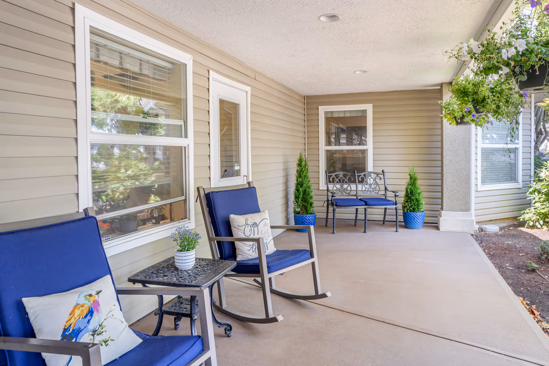 Covered outdoor patio area with beige siding walls, two blue cushioned rocking chairs with decorative pillows, a small black metal side table with a potted plant, a black metal bench with blue cushions, two potted plants in blue pots, and hanging flower baskets on the right side.