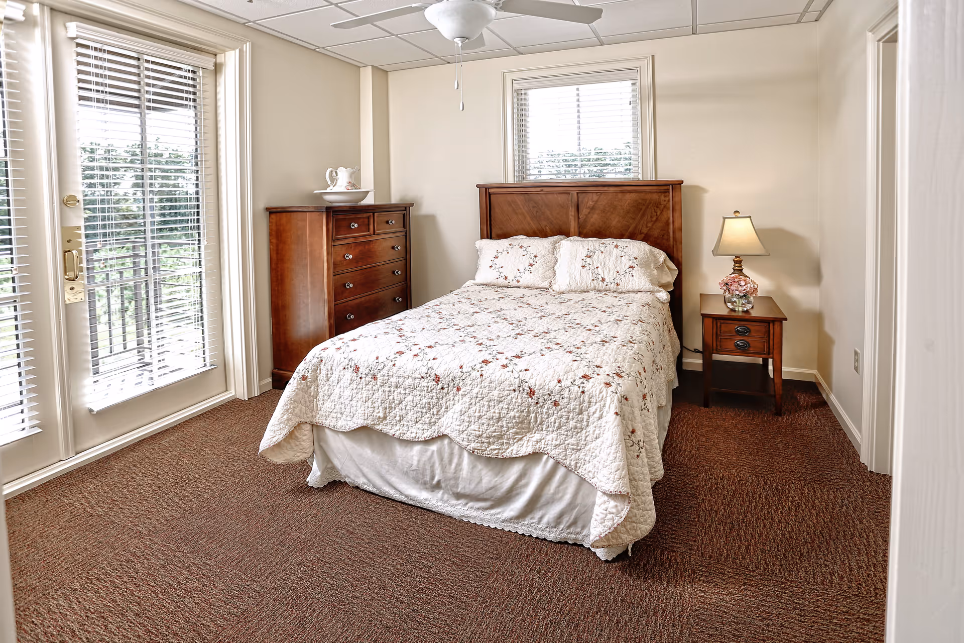 A cozy bedroom with a double bed covered in a white floral quilt. The room features a wooden headboard, a wooden chest of drawers with a decorative pitcher and bowl on top, and a wooden nightstand with a lamp and a small flower arrangement. There are two windows with blinds and a glass door leading to a balcony or outdoor area. The floor is carpeted in brown, and the ceiling has a white fan.