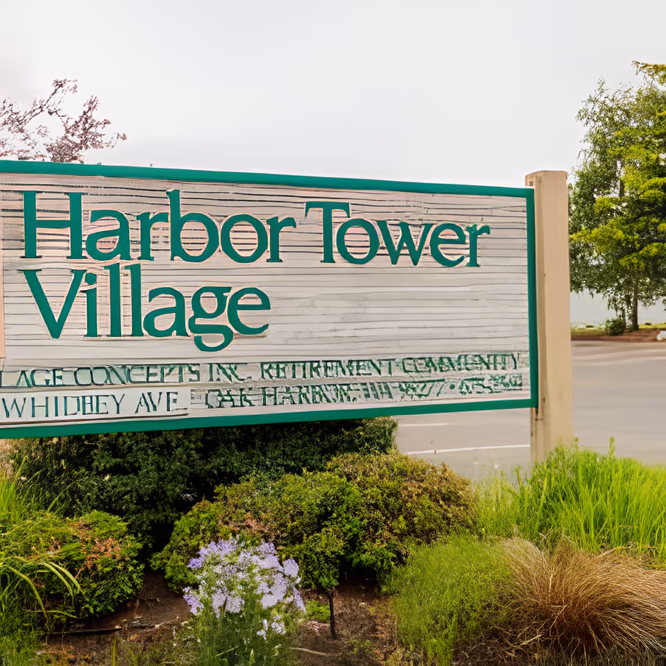 Outdoor image of a large wooden sign with green trim that reads 'Harbor Tower Village' surrounded by bushes and flowers, with a parking lot and trees in the background.