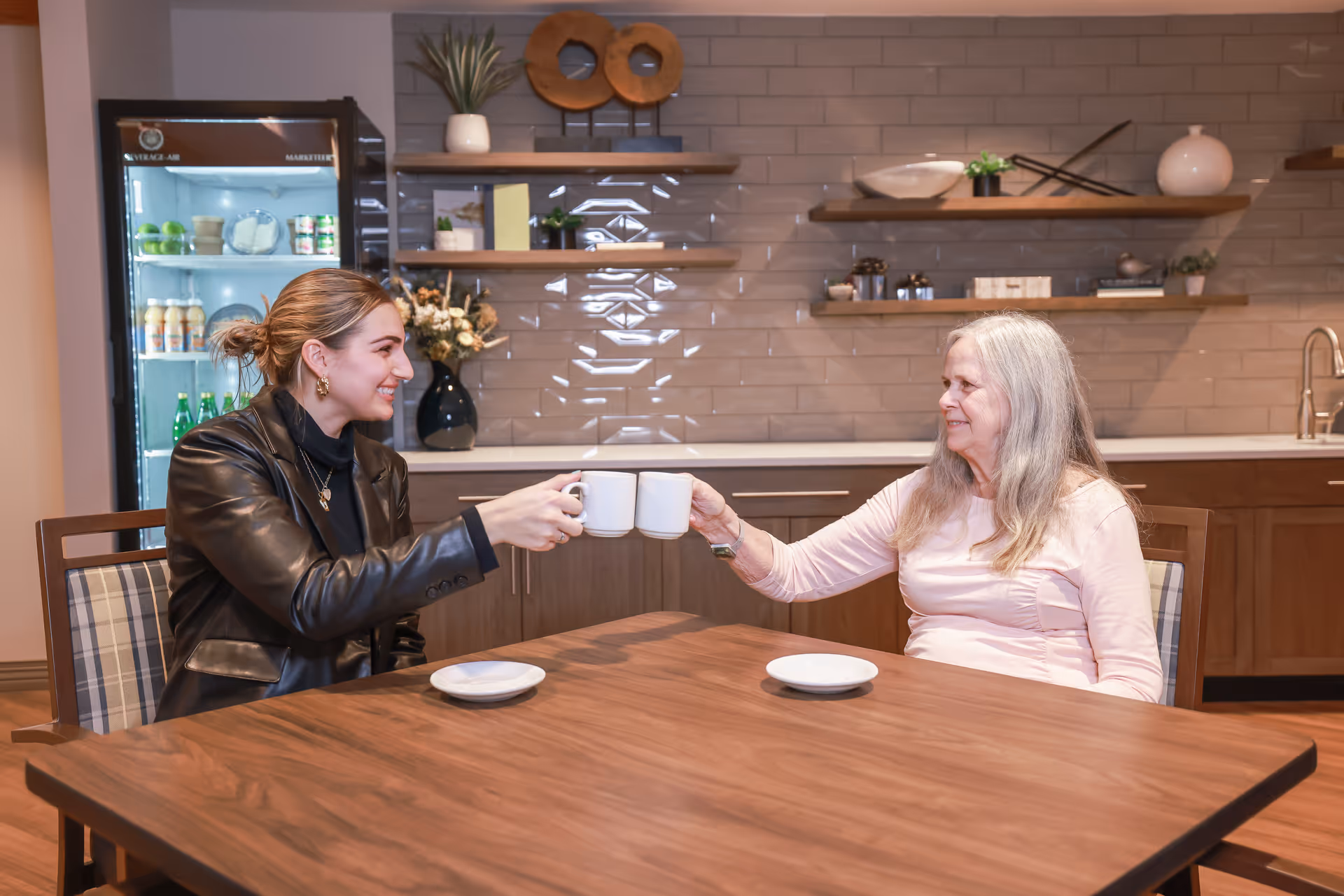 Two women sitting at a wooden table in a kitchen or dining area, smiling and clinking white coffee mugs. The background shows shelves with decorative items, a refrigerator with beverages, and a kitchen sink.