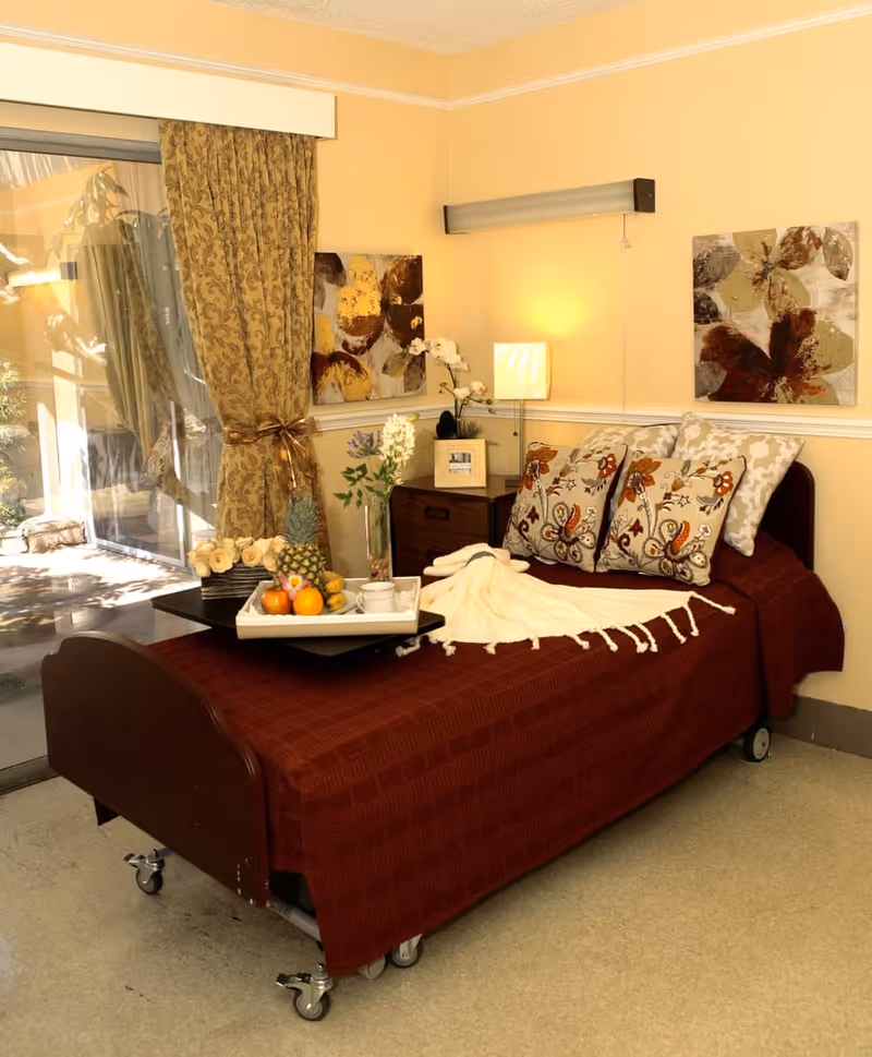 A cozy bedroom in a senior living facility with a single bed covered in a dark red blanket and decorative pillows. A white throw blanket is draped over the bed. A tray with fruit, a cup, and a vase with flowers is placed on the bed. The room has yellow walls, two floral paintings, a nightstand with a lamp, and a sliding glass door with patterned curtains leading outside.