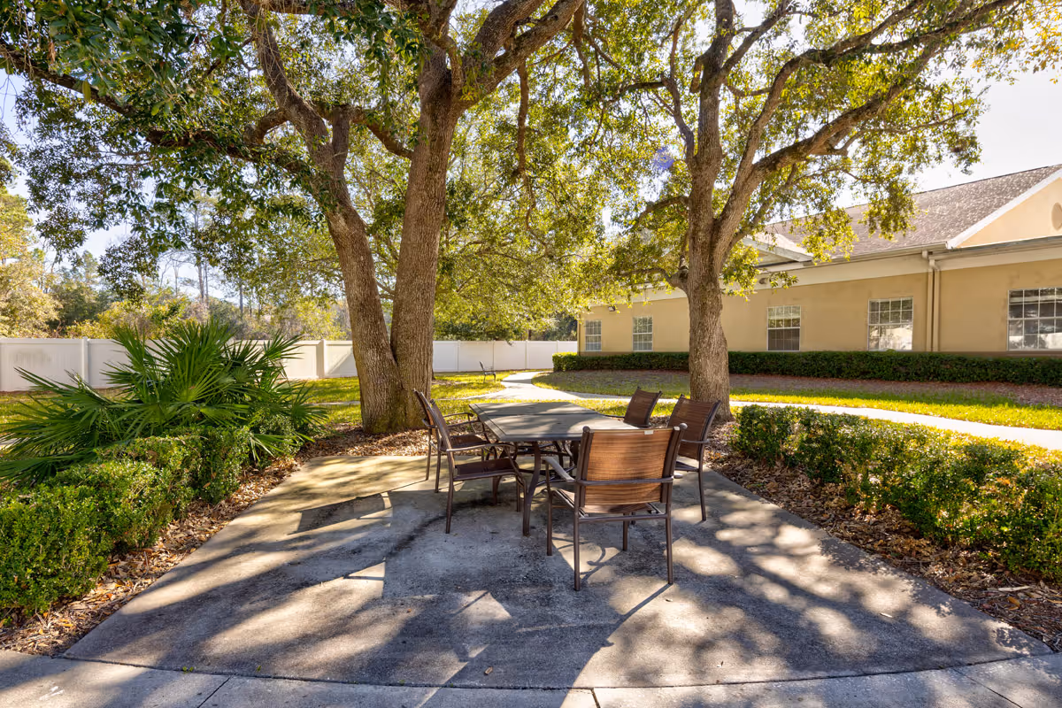Outdoor patio area with a rectangular table and six chairs under large trees providing shade. The patio is surrounded by green shrubs and plants, with a beige building and white fence in the background.