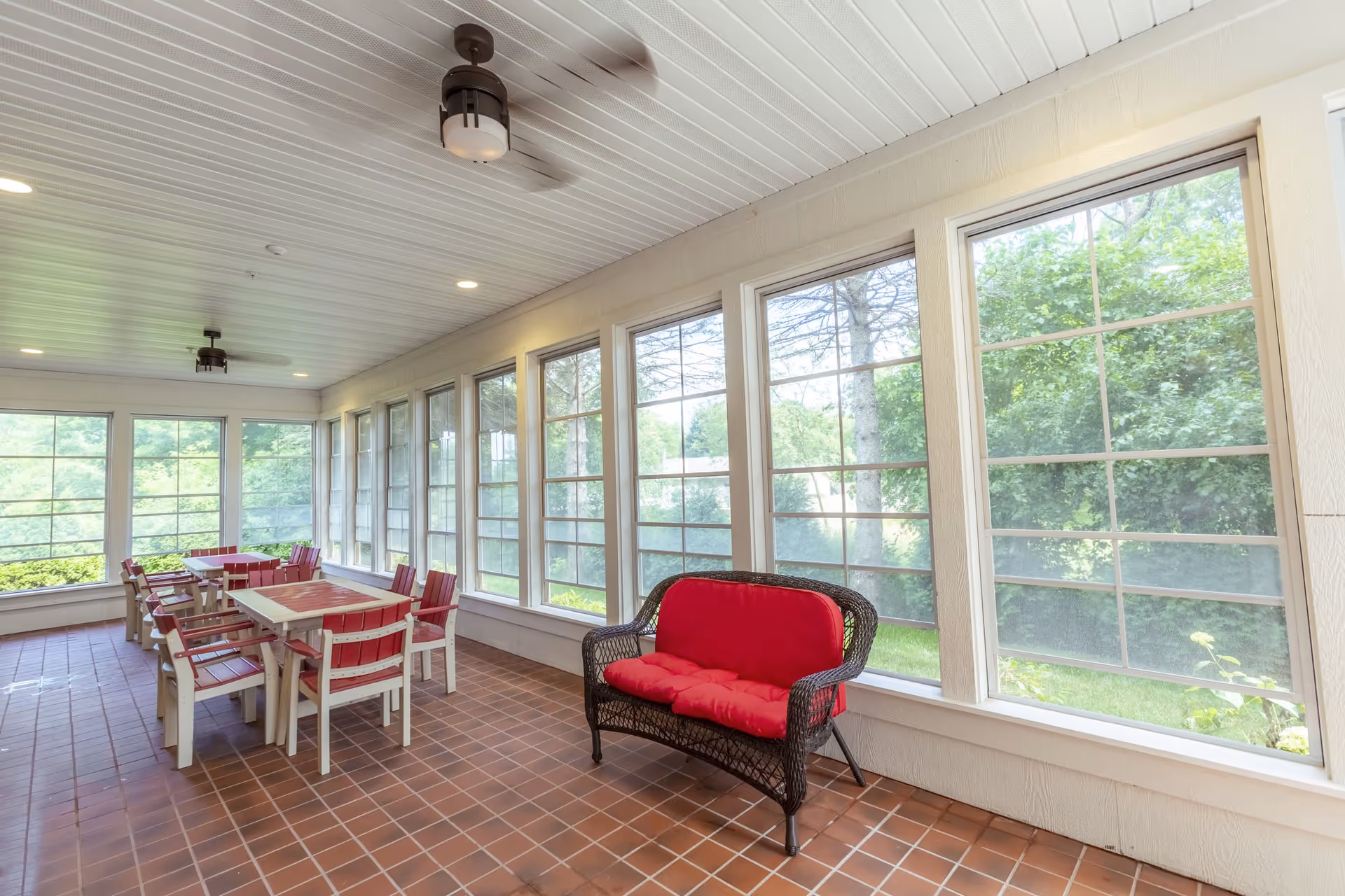 A bright enclosed porch area with large windows showing green trees outside. The room has a tiled floor, white paneled ceiling with ceiling fans, and several tables with red cushioned chairs. There is also a black wicker loveseat with red cushions near the windows.