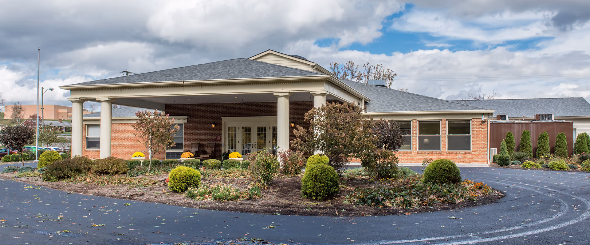 Exterior view of Lindside Healthcare Center showing a single-story brick building with a covered entrance supported by columns. The driveway curves around a landscaped area with bushes and small trees. The sky is partly cloudy.