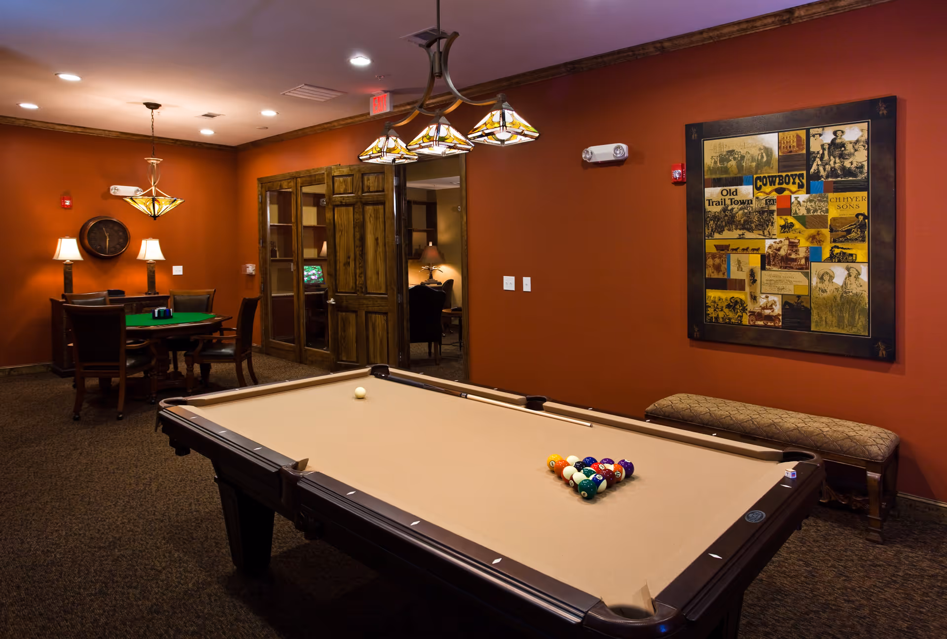 A recreational room with a pool table in the foreground, a bench against a red wall with a framed collage of vintage cowboy images, and a round table with chairs and lamps in the background. The room has warm lighting and wooden doors leading to another area.