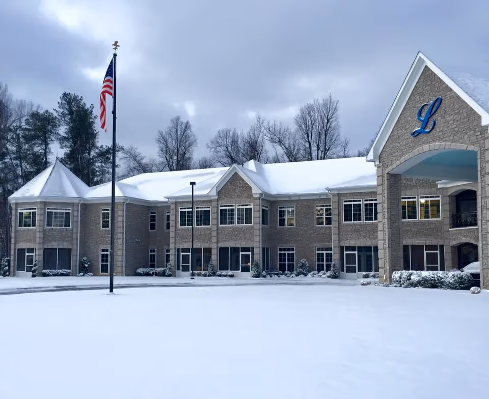 Snow-covered two-story brick building with a covered entrance and a blue 'L' emblem, an American flagpole in front, and bare trees behind.