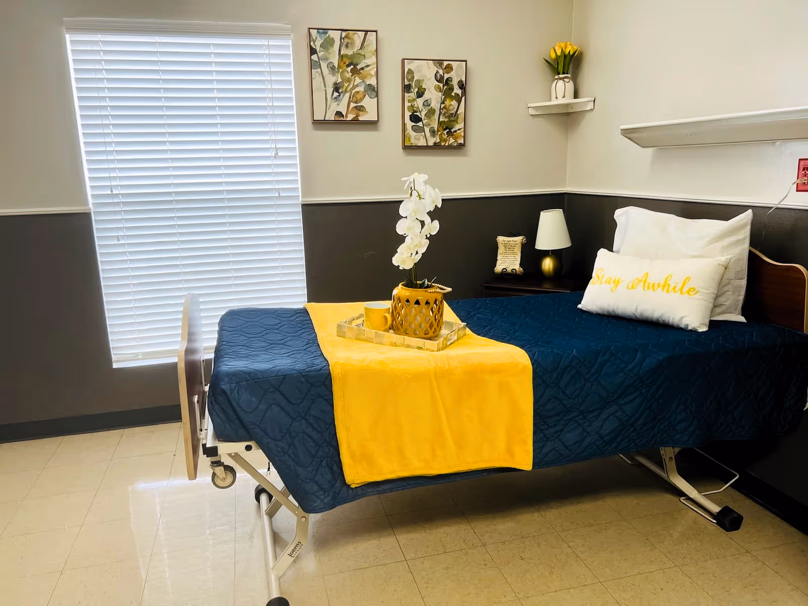 A neatly made single bed in a room with a blue quilt and a yellow throw blanket. On the bed is a tray with a yellow vase holding white flowers and a yellow mug. A white pillow with yellow text reading 'Stay Awhile' is placed at the head of the bed. The room has a window with closed white blinds, two framed botanical prints on the wall, a small nightstand with a lamp and decorative item, and a small shelf with a yellow flower vase.
