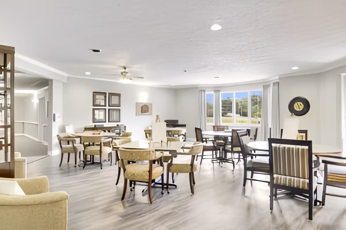 A bright and spacious dining room with multiple round tables and chairs arranged neatly. The room has large windows letting in natural light, light-colored walls, and a ceiling fan. There is a decorative wall piece and framed artwork on the walls.